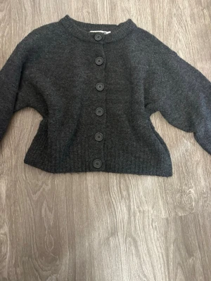 Mörkgrå stickad kofta med knappar - Mysig mörkgrå stickad kofta med stora mörkgrå knappar framtill. Koftan har rund halsringning, långa ärmar och ribbade muddar. Perfekt för lager-på-lager och enkel att matcha med jeans eller kjol. Koftan har också slitningar på sidorna, (PRISET GÅR ATT DISKUTERA KAN SÄNKA HUR MYCKET SOM HELST)!!
