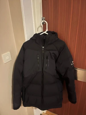 Svart pufferjacka från Sail Racing - Säljer en svart pufferjacka från Sail Racing med huva och två vertikala bröstfickor med dragkedja. Jackan har en sportig look och är perfekt för kalla vinterdagar. Tillverkad i slitstarkt material och designad för att hålla värmen. Snygg logga på ärmen.