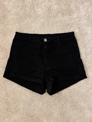 Svarta denimshorts  - Svarta shorts i denim med hög midja och klassisk femficksdesign. De har uppvikta benslut och stängs med knapp och dragkedja framtill. 