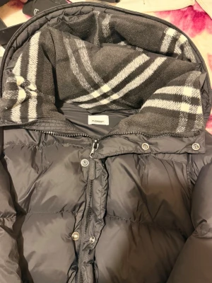 Svart pufferjacka från Burberry - Säljer en svart pufferjacka från Burberry med klassiskt rutigt foder i huvan. Jackan har dragkedja och tryckknappar framtill samt logga på ärmen. Perfekt för kalla vinterdagar och riktigt snygg streetstil.