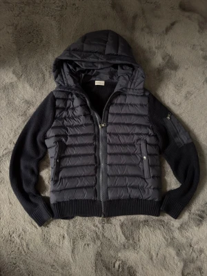 Mörkblå pufferjacka med huva från Moncler - Snygg mörkblå pufferjacka från Moncler med stickade ärmar och huva. Jackan har dragkedja framtill, två fickor med tryckknappar och en extra ficka på ärmen. Perfekt för kalla vinterdagar och riktigt trendig look.