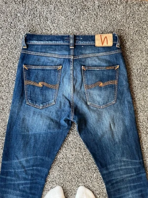 Blå jeans från Nudie Jeans - Snygga blå jeans från Nudie Jeans i modell lean Dean med klassisk femficksdesign och orangea sömmar på bakfickorna. Jeansen har en rak passform och är tillverkade i denim med en lätt tvättad look. Perfekta för en avslappnad stil. Storleken är 31/30 men sitter mer som 30/30