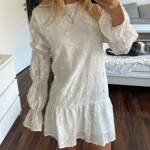 Vit blommig kortklänning med öppen rygg - Supersöt vit kortklänning med broderade blommor, volang nertill och puffiga långärmar med rosett vid handleden. Klänningen är från NA-KD, nypris 699kr