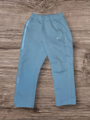 Blå mjukisbyxor från Nike NOCTA - Snygga ljusblå mjukisbyxor från Nike NOCTA med elastisk midja och raka ben. Byxorna har en diskret Nike-logga framtill och NOCTA-tryck på benet. Tillverkade i mjukt material som passar perfekt för chill eller träning. Har lite smy stygn som inte har gått upp men har blivit lite stretch out men ska inte vara ett problem. Hade lite små fläckar innan men har tvättat bort det mesta