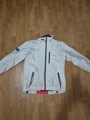 Vit jacka från Helly Hansen - Vit vindjacka från Helly Hansen med svarta detaljer och röda dragkedjor. Strl S, fråga vid fundering 