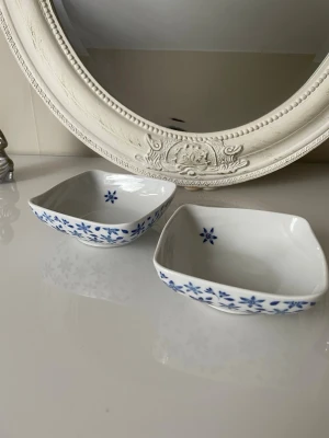 Fyrklövern Lise Finckenhagen – 2 st porslinsskålar blå/vita - Två fyrkantiga skålar i vitt porslin med blå blommönster längs kanterna och en blå blomdetalj i botten. Perfekta för snacks eller små tillbehör. Klassisk och stilren design som passar i de flesta hem.