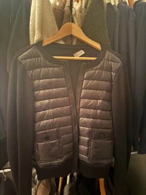 Svart cardiganjacka från Moncler - Snygg svart cardiganjacka från Moncler med quiltade partier framtill och två fickor med knapp. Jackan har rund halsringning, dragkedja och stickade ärmar i mjukt material. Perfekt för lager-på-lager och streetstyle.