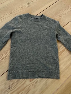 Grå stickad tröja från GANT - Mysig grå stickad tröja från GANT med rund halsringning och diskret broderad logga på bröstet. Tröjan har armbågslappar i en kontrasterande färg. Nypris 2500 kr Tillverkad i ull