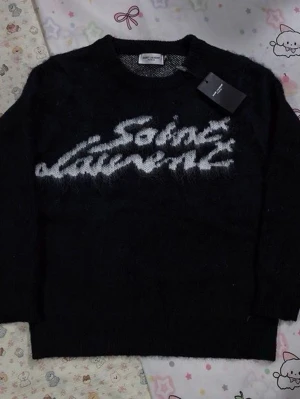 Saint laurent tröja - Snygg svart stickad tröja från Saint Laurent med vit logotyp i graffiti-stil över bröstet. Tröjan har rund halsringning och långa ärmar. Perfekt för dig som gillar exklusiva och trendiga plagg.