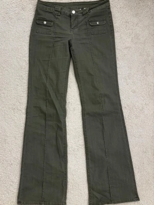 Olivgröna bootcut jeans H&M - Snygga olivgröna jeans med bootcut passform och två framfickor med lock och knappdetaljer. Jeansen har klassisk knappgylf och bälteshällor. Perfekta för dig som gillar en retroinspirerad look och vill ha något utöver det vanliga. (säljs inte längre)