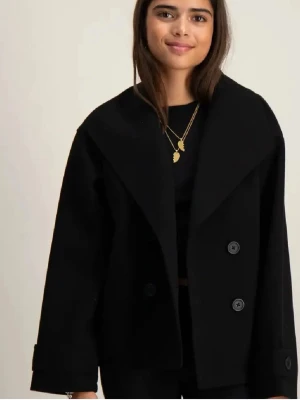 Svart oversized kappa med bred krage - Stilren svart kappa med bred krage och dubbelknäppning framtill. Modellen är oversized med raka ärmar och klassiska knappar. Perfekt för dig som gillar minimalistisk och modern stil. Materialet ser ut att vara ullblandning.storlek 158/164 motsvarar xs 