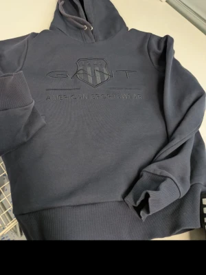 Mörkblå hoodie från GANT - Snygg mörkblå hoodie från GANT med broderad logga och texten 'American Sportswear' på bröstet. Klassisk huva med dragsko och ribbade muddar vid ärmar och midja. Perfekt för en avslappnad och sportig stil.