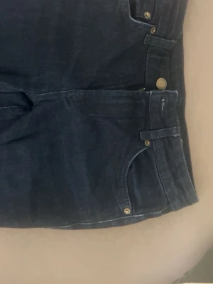 Mörkblå bootcut jeans - Säljer ett par mörkblå bootcut jeans med klassisk femficksdesign och snygga mässingsfärgade nitar. Jeansen har hög midja och raka ben som går ut lite nedtill. Perfekta till sneakers eller boots.