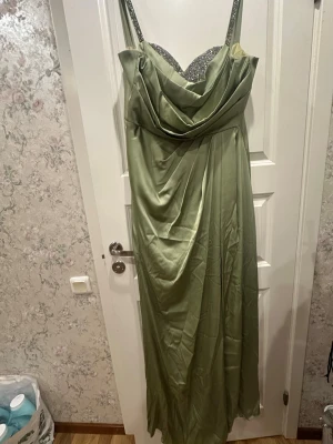 Grön långklänning med glitterdetaljer - Elegant långklänning i ljusgrönt satinliknande material med glittriga axelband och bystparti. Draperad framtill för en snygg siluett och smala axelband. Perfekt för bal eller festliga tillfällen.