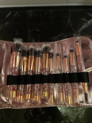 Glittrigt sminkborstset i roséguld - Ett set med tio sminkborstar med glittriga handtag i roséguld och transparent plast. Borstarna kommer i ett snyggt fodral i metallicrosa. Perfekt för att fixa hela sminkningen, från bas till ögonbryn. Borsthuvudena är täta och mjuka, och varje borste har ett eget fack i fodralet.