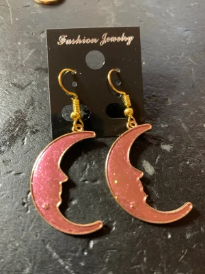 Lila måneörhängen med ansiktsprofil - Unika örhängen i form av lila halvmånar med ansiktsprofil och små stjärndetaljer. De har guldfärgade krokar och en glansig finish. Perfekta för dig som gillar statement-smycken med en magisk touch.