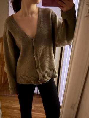 Beige kofta från Zara med v-ringning - Mysig beige kofta från Zara med djup v-ringning och knäppning framtill. Koftan har långa ärmar och breda ribbade muddar vid ärmslut och nederkant. Perfekt att slänga på sig över en t-shirt eller linne.