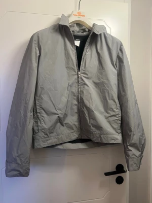 Filippa K Ljusgrå vindjacka med dragkedja - Stilren ljusgrå vindjacka med klassisk krage och hel dragkedja framtill. Jackan har raka ärmar med breda muddar och är tillverkad i ett tunt, vindtätt material. Perfekt för blåsiga dagar och enkel att matcha med olika outfits.