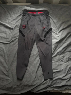 Grå PSG träningsbyxor från Nike - Snygga grå träningsbyxor från Nike med PSG-logga och röd Nike swoosh på framsidan. Byxorna har smal passform, elastisk midja och svarta detaljer längs sidorna. Perfekta för fotbollsträning eller chill. Materialet är troligtvis polyester för maximal komfort.