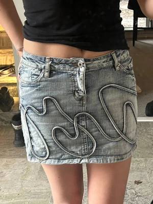 Ljusblå denim minikjol med detaljer - Trendig ljusblå jeanskjol i kort modell med unika sydda vågiga detaljer framtill och bak. Kjolen har klassiska fickor, bälteshällor och nitar på bakfickan. Perfekt för dig som vill sticka ut med en cool streetstyle-look.