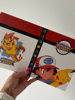 Pokémon Brilliant Stars samlarpärm - Pokémon samlarpärm med färgglada motiv av Pikachu, Ash och Brilliant Stars-logga. Pärmen är röd, vit och svart med plastfickor för kortförvaring. Perfekt för att organisera och visa upp dina Pokémonkort. Plats för 240 kort 