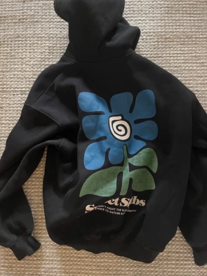 Svart hoodie med tryck - Säljer en svart hoodie från Weekday med ett coolt tryck på ryggen, en stor blå blomma och gröna blad. Hoodien har huva och ribbade muddar vid ärmslut och nederkant. Materialet är mjukt och skönt, perfekt för en avslappnad stil. Hoddien är lite oversized och skön. Framsidan är endast svart. 