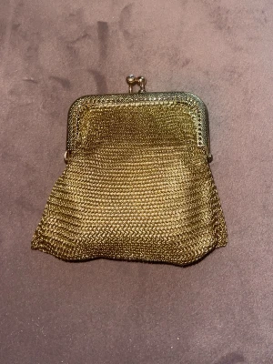 Liten guldig metallplånbok - Säljer en liten vintageinspirerad plånbok i guldig metall med nätstruktur och klassiskt bygelspänne. Perfekt för mynt eller småsaker och har en unik retrostil som sticker ut i mängden.