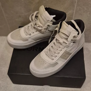 Beige höga sneakers från Björn Borg strlk 37 - Snygga höga sneakers från Björn Borg i beige och ljusgrått. Skorna har klassisk snörning, perforerade detaljer på tån och vadderad plös med logga. Ovandelen är i syntetmaterial och mockaimitation, med svart innerfoder för extra kontrast. Dessa växtes ur innan de hann användas. Nya i kartong.