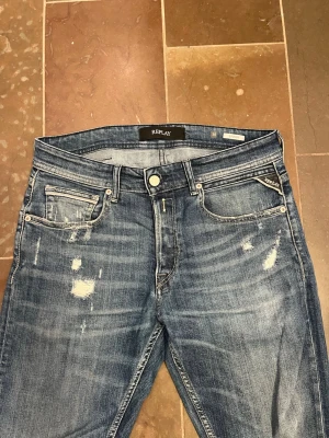 Blå slitna jeans från Replay - Säljer ett par blå jeans från Replay med snygga slitningar och klassisk femficksmodell. Jeansen har en rak passform och detaljerade sömmar på bakfickan. Materialet är mjukt denim med en tvättad look som ger en avslappnad vibe.