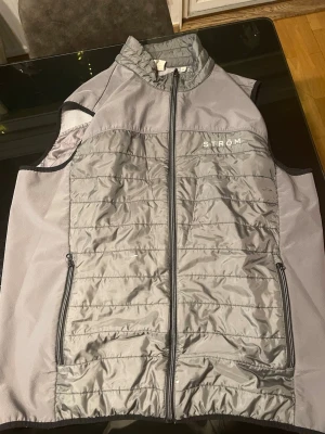 Grå dunväst från STRÖM - Snygg grå dunväst från STRÖM med quiltad framsida och baksida, hel dragkedja och två dragkedjefickor. Västen har hög krage och är tillverkad i ett lätt, glansigt material framtill och bak, med mjukare tyg på sidorna. Perfekt för lager-på-lager-stil.