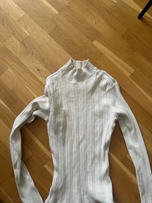 Vit ribbad stickad polotröja - Säljer en vit ribbad stickad tröja med hög krage och långa ärmar. Tröjan har en smal passform och vertikala ränder i stickningen som ger en snygg struktur. Perfekt att styla med jeans eller kjol för en clean look.