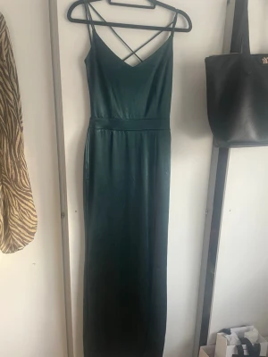 Mörkgrön balklänning med slits och spets - Snygg mörkgrön balklänning i satinliknande material med smala axelband som korsas i ryggen, även spets i ryggen och en slits på sidan. Använd 2 gånger