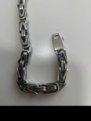 Tungt kedjearmband i silverfärg  - Säljer ett stilrent armband i silverfärgad metall med robust, geometrisk kedjedesign. Länkarna är breda och har en unik, vinklad form som ger ett coolt och modernt intryck. Perfekt accessoar för dig som gillar statement-smycken och en edgy look.