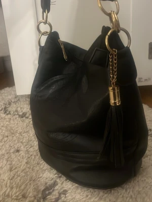 Svart bucket bag med gulddetaljer - Stilren svart bucket bag i slätt med snygga guldfärgade metalldetaljer och dekorativ tofs. Väskan har en bred axelrem och dragkedja baktill för extra förvaring. Perfekt för dig som gillar en trendig och rymlig handväska.
