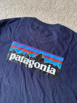 Patagonia mörkblå t-shirt, storlek M - T-shirt från Patagonia med kort ärm och rund halsringning. Tröjan har en liten logotyp tryckt på vänster bröst och en större logotyp tryckt på ryggen.  T-shirten är i nyskick  Herrmodell 