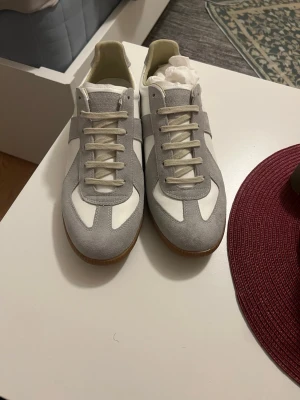 Maison margiela  - Snygga sneakers i vitt och ljusgrått med ovandel i mocka och skinn. Klassisk design med snörning och platt sula i gummi. Perfekta för dig som gillar stilrena och tidlösa sneakers. Märket Maison Margiela väldigt fina o sköna skor passar perfekt inför kommande årstider våren och sommaren väldigt stiliga och passar med allt. PRISET KAN DISKUTERAS:) Kvitto finns!!