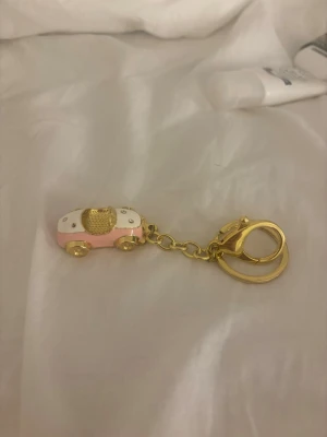 Nyckelring med bil i rosa och guld - Nyckelring med en liten bil i rosa, vitt och guld. Bilen har guldfärgade hjul, detaljer och glittriga stenar på ovansidan. Kedja och ring i guldfärgad metall. Perfekt accessoar till nycklar eller väska.