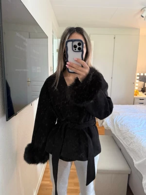 Svart pälsjacka med knytband - Säljer en svart pälsjacka med breda ärmar och fluffiga pälsdetaljer vid ärmslut och krage. Jackan har ett knytband i midjan som ger en snygg siluett. Perfekt för att hålla sig varm och stilren under kalla dagar. Använd endast en gång men inte riktigt min stil. 
