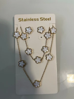 Guldhalsband med vita blommor - Snyggt halsband i guldton med vita blommor i pärlemor-look. Blommorna är jämnt placerade längs hela kedjan och ger ett elegant och trendigt intryck. Halsbandet är tillverkat i rostfritt stål och passar perfekt för dig som gillar unika accessoarer.