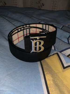 Svart Burberry bälte med silverdetalj - Snyggt svart bälte från Burberry med klassiskt rutigt innerfoder och en stor silvrig B-spänne framtill. Bältet är gjort i präglat läder med diskret mönster och har en exklusiv känsla. Perfekt accessoar för att lyfta din outfit.