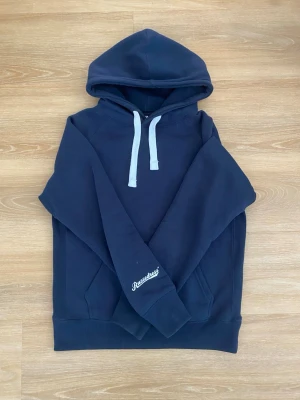 Mörkblå hoodie från Russemerch  - Mörkblå hoodie från Russedress med vit dragsko i huvan och logga tryckt på ärmen. Klassisk känguruficka framtill och ribbade muddar vid ärmslut och nederkant. Perfekt för en avslappnad och stilren look.
