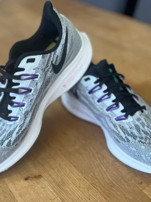 Nike grå sneakers med svart swoosh - Snygga sneakers från Nike i grått med svart swoosh på sidan. Ovandelen är i meshmaterial med stickat mönster och svarta samt lila detaljer på snörningen. Sulan är vit med färgglada inslag undertill. Perfekta för en sportig och trendig look.