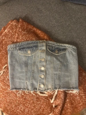 Ljusblå denim tubtopp med knappar - Trendig ljusblå tubtopp i denim med fransig nederkant och snygga silverfärgade knappar framtill. Den är ganska kort som man kan se på bilden men väldigt snygg! Använd enstaka gånger.