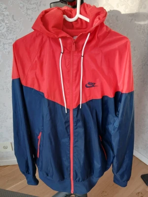 Nike vindjacka röd och marinblå - Nike vindjacka i rött och marinblått med dragkedja, huva och vita dragsnören. Jackan har meshfoder, två fickor med dragkedja och klassisk Nike-logga på bröstet. Perfekt för blåsiga dagar och sportig stil.