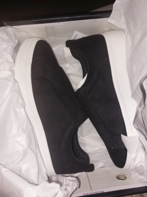 Svarta slip-on sneakers med vit sula - Stilrena svarta slip-on sneakers med bred resår över vristen och vit, chunky sula. Ovandelen är i textilmaterial som ger en clean look och skorna är lätta att ta på. Perfekta för dig som gillar minimalistisk stil och vill ha något enkelt men snyggt.