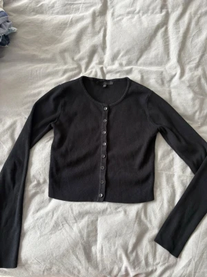 Svart cropped kofta med knappar - Svart, ribbstickad cropped kofta från Monki med rund halsringning och knäppning framtill. Långa ärmar och tajt passform, perfekt att styla över linne eller t-shirt. Enkel och stilren design som funkar till många outfits.