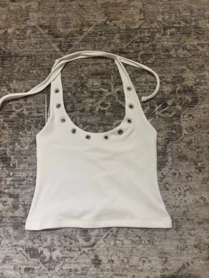 Vit ribbad linnetopp från Edited - Snygg vit ribbad linnetopp från Edited med silverfärgade öljetter runt den djupa urringningen och knytband i nacken. Perfekt för dig som gillar trendiga detaljer och en clean look.