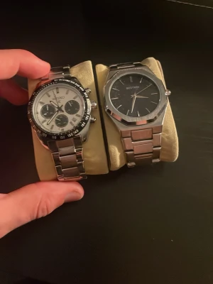 Seiko och Bestwin herrklockor i stål - Två stilrena herrklockor med armband i silverfärgat stål. Seiko-klockan har vit urtavla med svarta detaljer och tre subdials, medan Bestwin har en svart urtavla och minimalistisk design. Båda har robusta länkar och klassisk form.