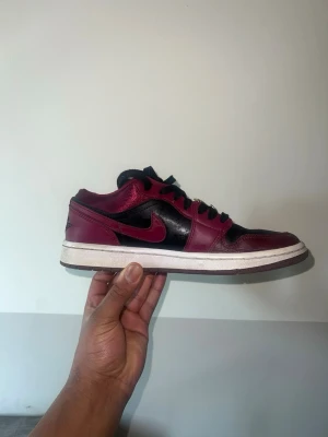 Nike Air Jordan 1 Low svart/röd - Snygga Nike Air Jordan 1 Low sneakers i svart och mörkrött läder. Klassisk siluett med svart swoosh och röda detaljer, vit mellansula och svart yttersula. Perfekta för dig som gillar streetwear och vill sticka ut med en ikonisk modell.