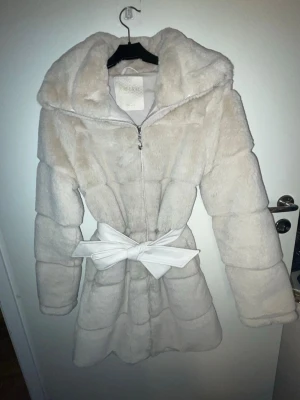 Beige pälsjacka med huva och rosett - Supermjuk beige pälsjacka från Missu med stor huva och dragkedja framtill. Jackan har en markerad midja med en bred vit rosett som knyts framtill. Perfekt för kalla vinterdagar och ger en lyxig känsla till din outfit.